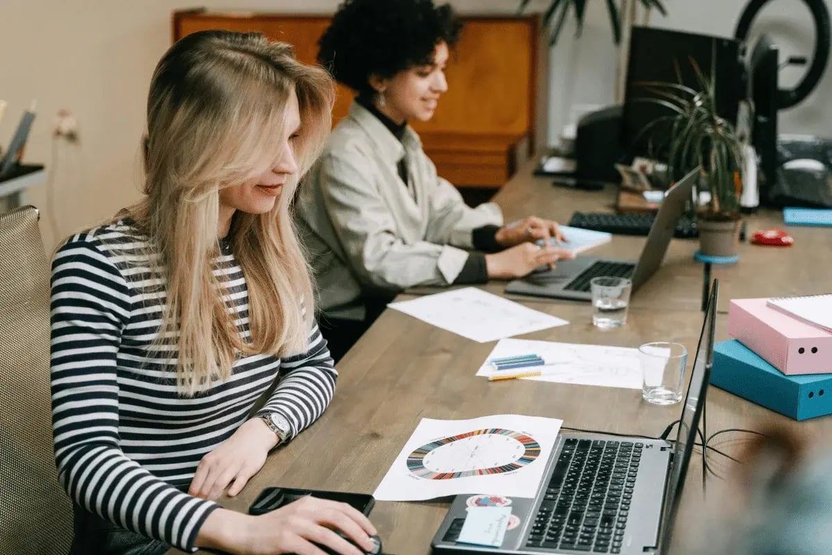 Two individuals work at a wooden desk, focused on laptops surrounded by documents, stationery, and a plant.