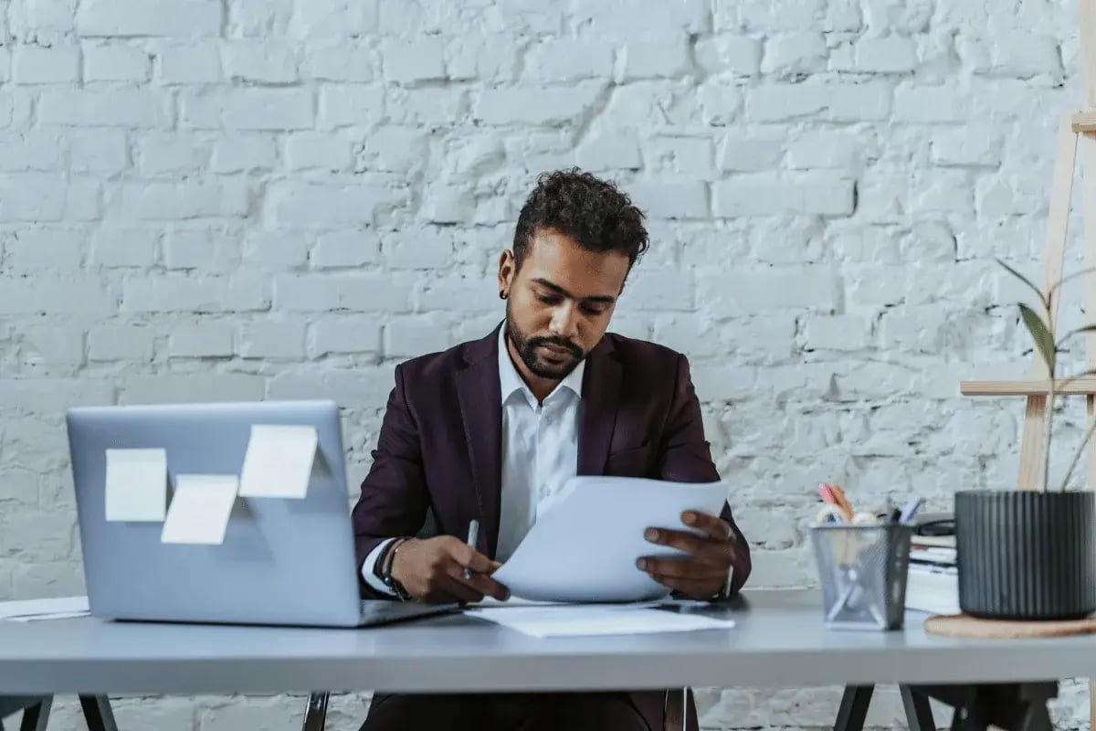 A man in a dark suit sits at a desk with a laptop, reading and writing on papers. Sticky notes are on the laptop, and a plant is in the background.