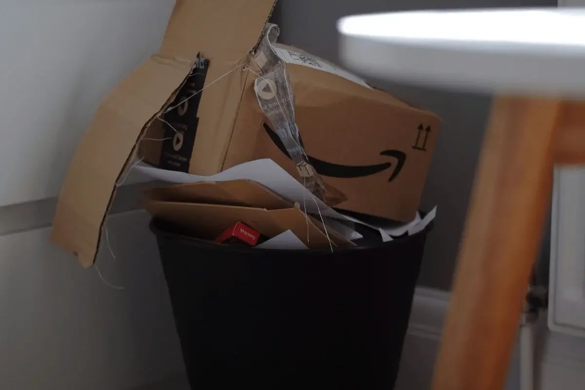 A black trash bin filled with discarded papers and an Amazon cardboard box, partially crumpled and leaning against the bin.