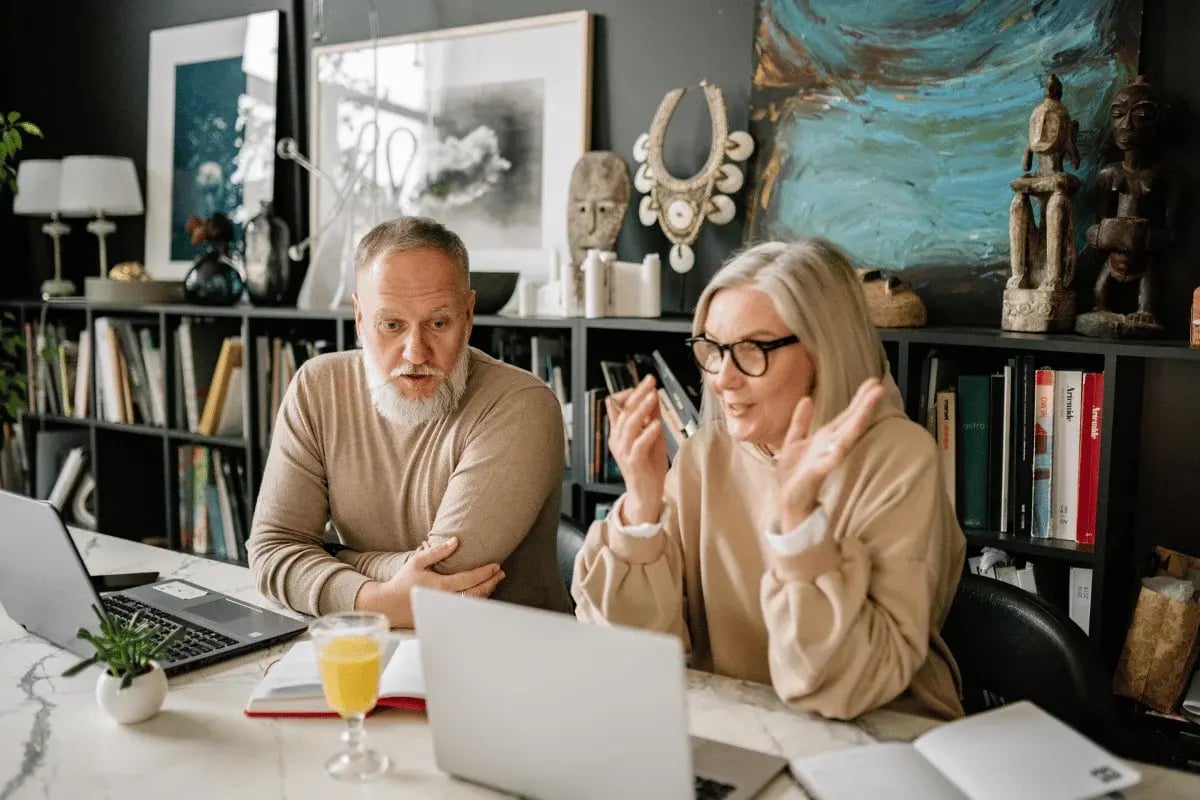 A man and woman engaged in a discussion sit at a desk with laptops, books, and a glass of orange juice. Art and sculptures adorn the shelf behind them.