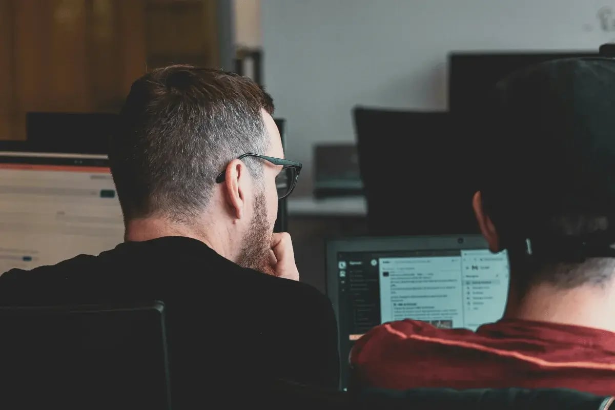 Two people are sitting in an office, focused on computer screens displaying text.